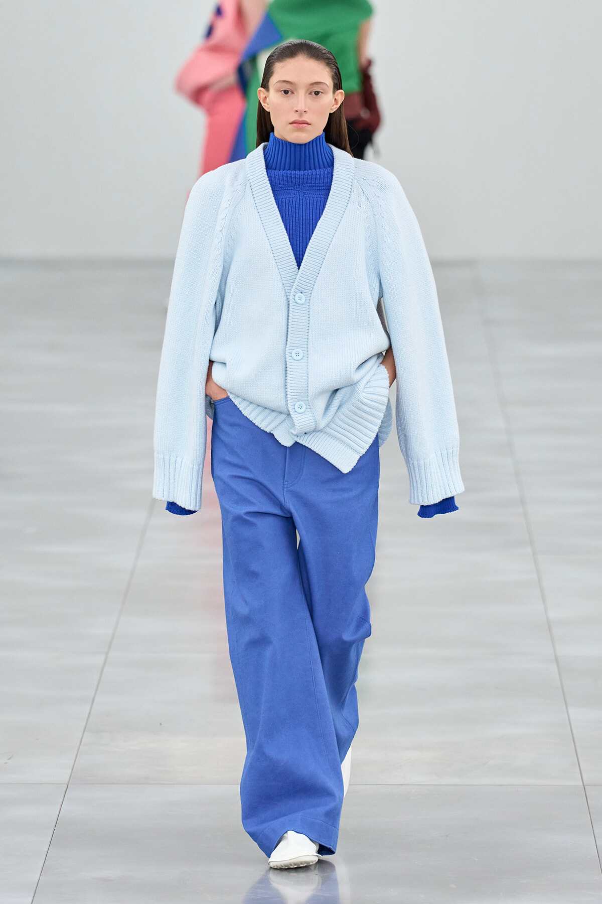 Model walks the runway in a pale blue oversized cardigan over a bright blue turtleneck and matching wide-leg pants, hands in pockets.