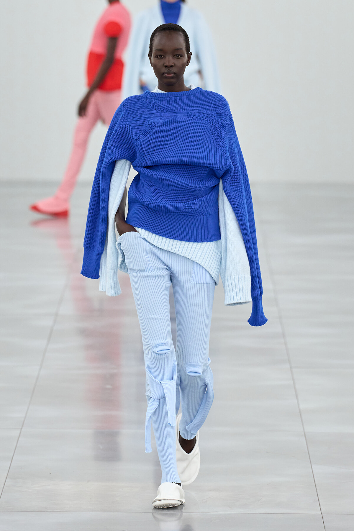 Model walking a fashion runway in a cobalt blue oversized knit sweater layered over light blue ribbed pants with white sneakers.