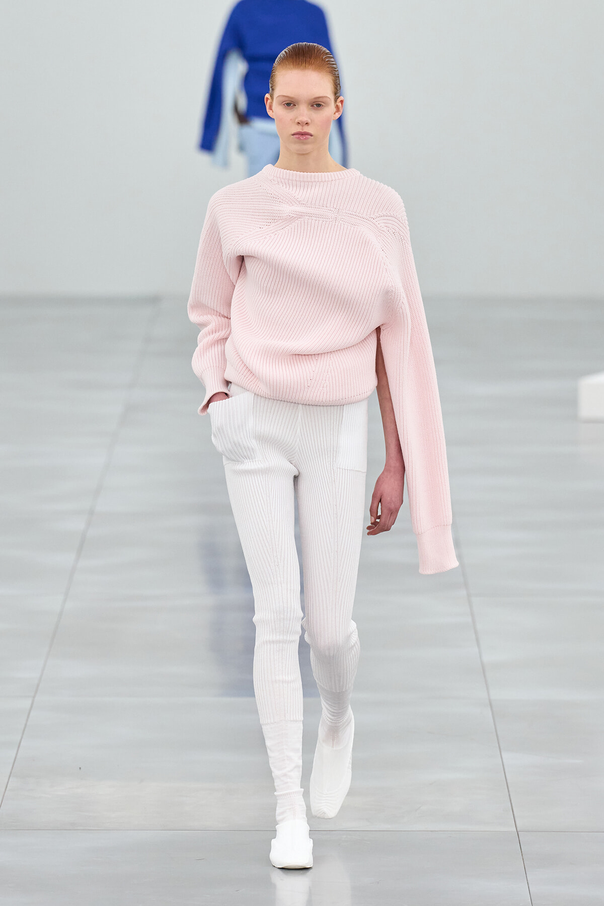 Runway model wearing a soft pink oversized knit sweater with white ribbed pants on a minimalist catwalk.