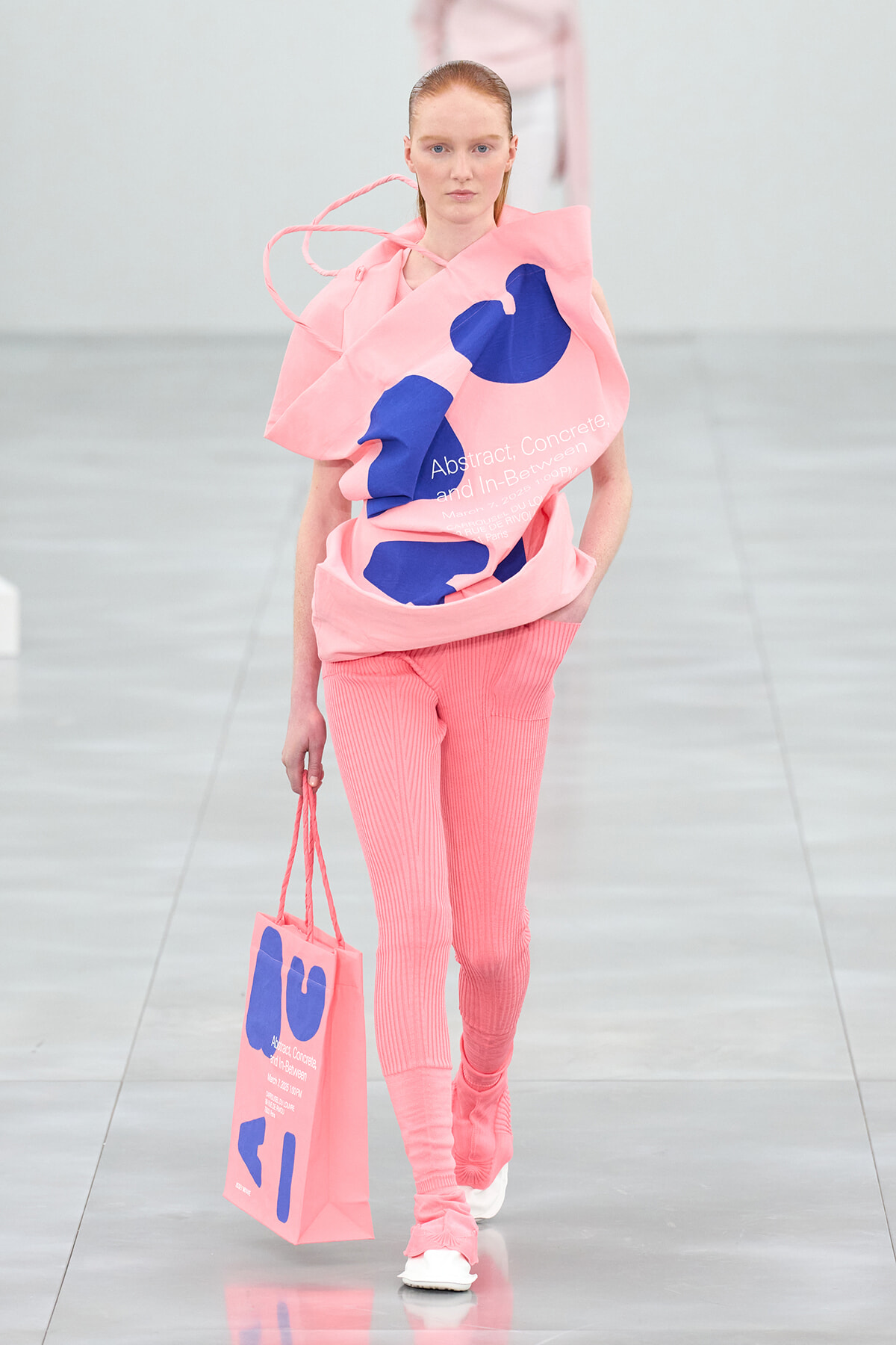 Model walking a fashion show wearing a pink oversized top with blue abstract shapes and matching pink ribbed pants, carrying a pink tote bag.