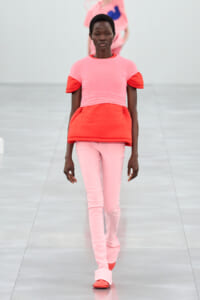 Model walking a runway in a pink and red layered outfit, pink top with red sleeves and midsection over pale pink pants.