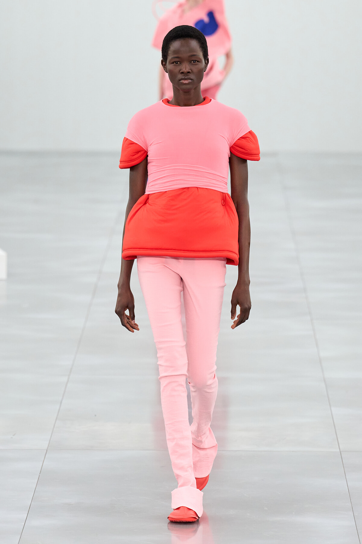 Model walking a runway in a pink and red layered outfit, pink top with red sleeves and midsection over pale pink pants.