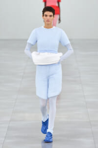 Model walking the runway in a pale blue ensemble with a white hip bag, white tights, and blue shoes on a gray catwalk
