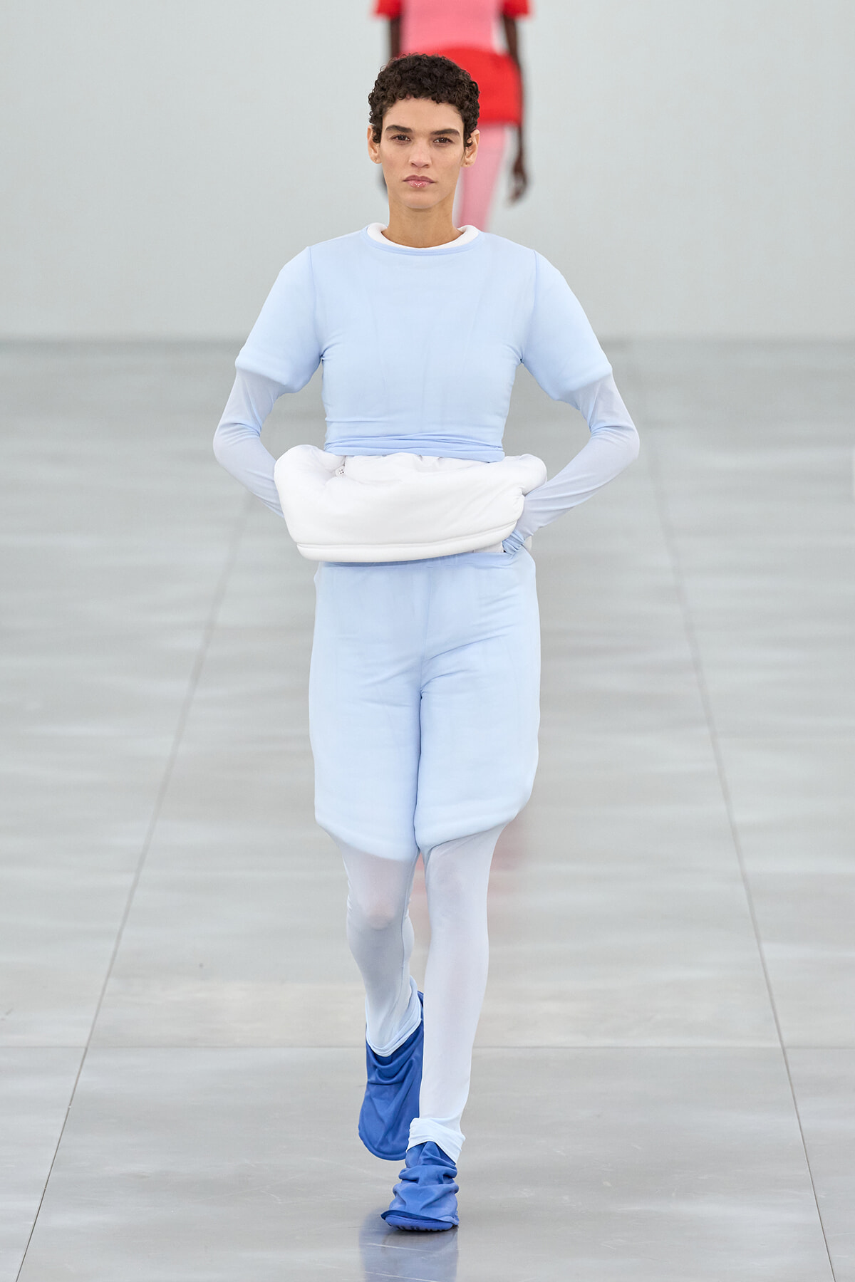 Model walking the runway in a pale blue ensemble with a white hip bag, white tights, and blue shoes on a gray catwalk
