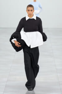 Model walks a runway wearing a black top with a white oversized collar and a large white padded wrap over black pants, holding extra padding pieces under her arm.