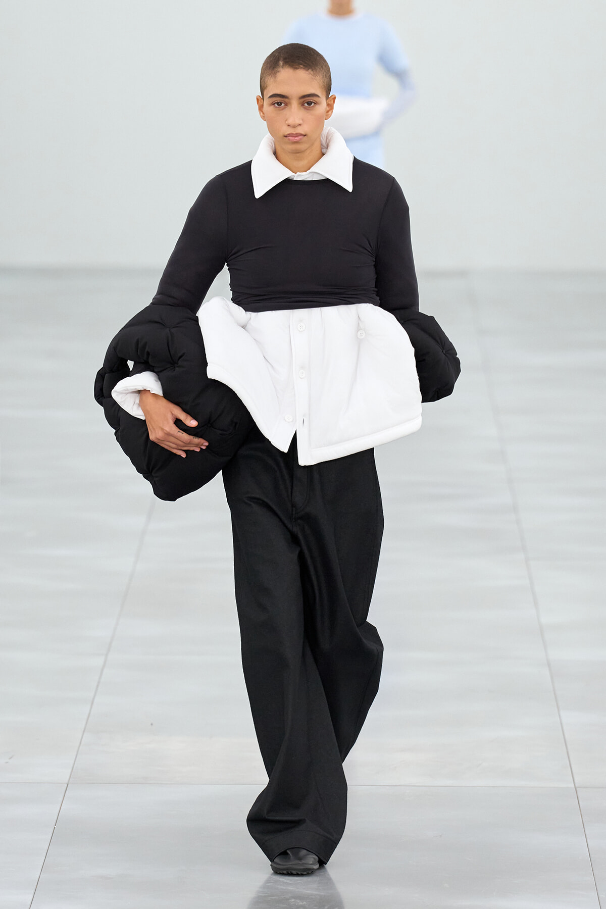 Model walks a runway wearing a black top with a white oversized collar and a large white padded wrap over black pants, holding extra padding pieces under her arm.
