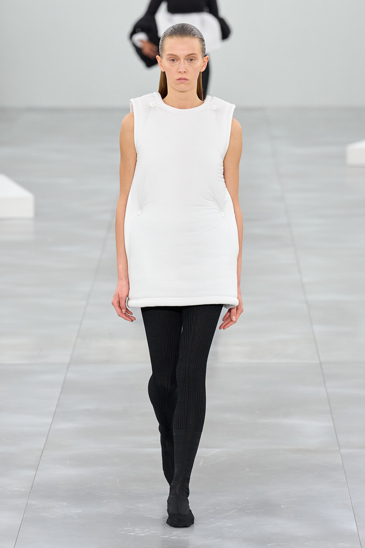 Model walking the runway in a white sleeveless tunic with button accents and black ribbed tights.