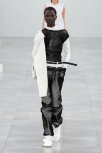Model walks a runway in a monochrome black-and-white outfit: sheer black top with white undershirt, white belt, distressed black pants, and a white coat draped over the left shoulder. White boots complete the look.