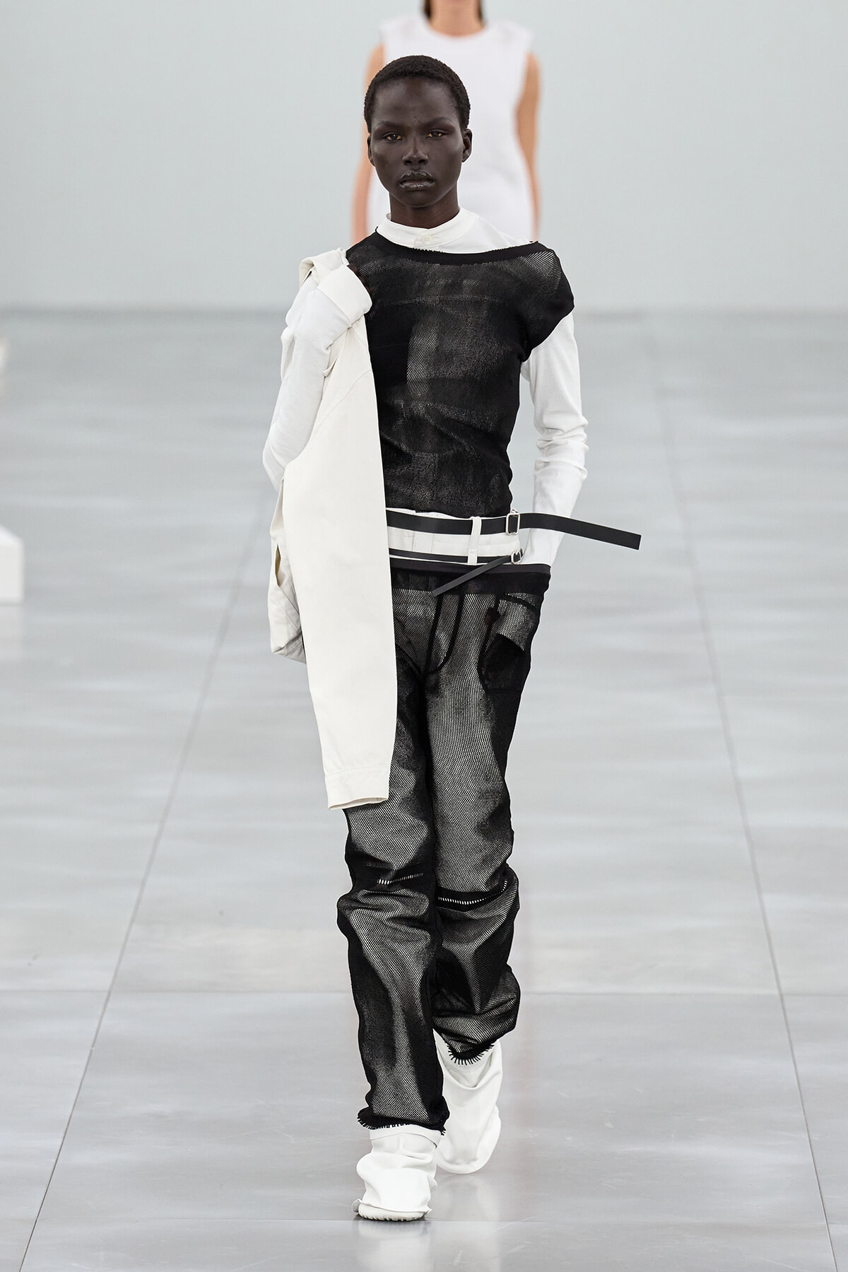 Model walks a runway in a monochrome black-and-white outfit: sheer black top with white undershirt, white belt, distressed black pants, and a white coat draped over the left shoulder. White boots complete the look.