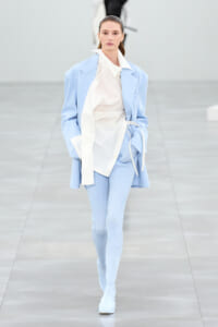 Runway model wearing a pale blue oversized blazer over a white shirt and light-blue ribbed leggings, walking the catwalk.