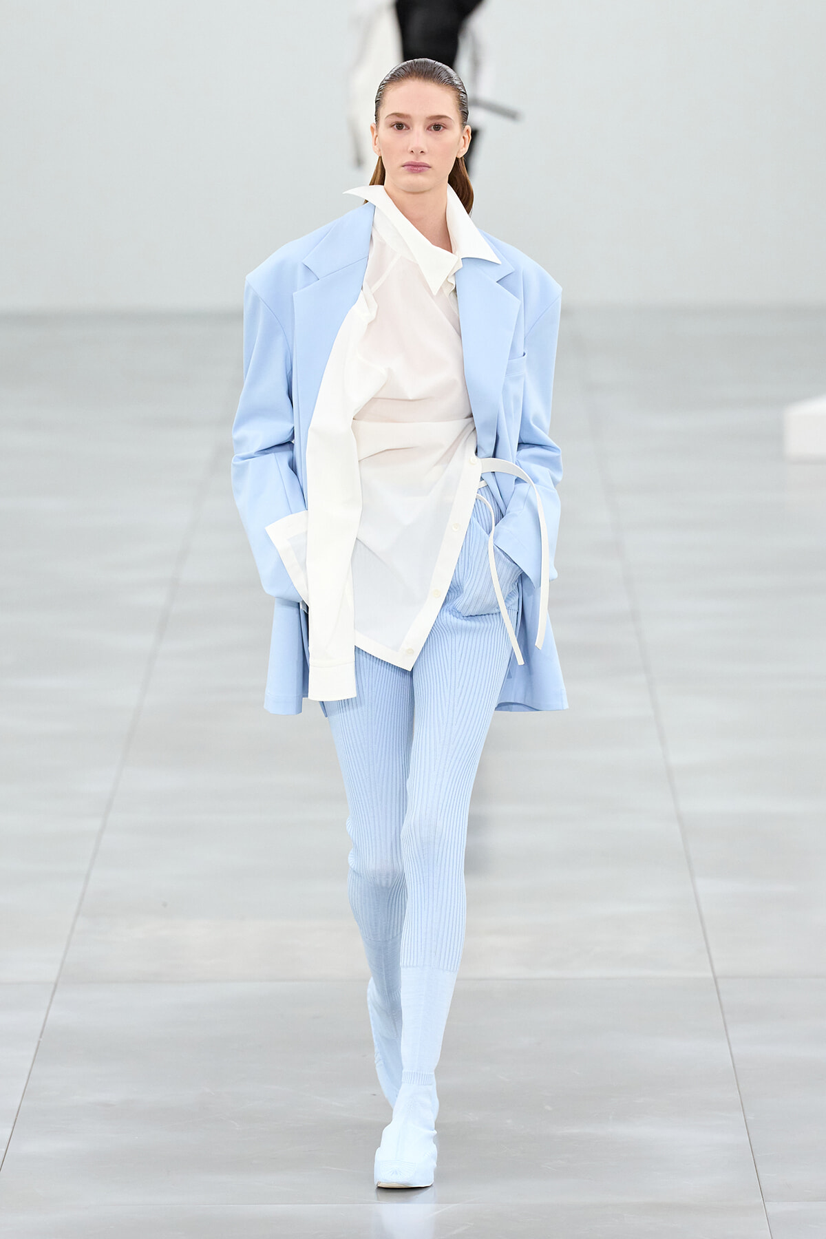 Runway model wearing a pale blue oversized blazer over a white shirt and light-blue ribbed leggings, walking the catwalk.