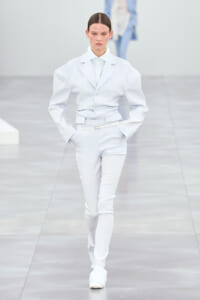 Model on the runway wearing a pale blue tailored suit with a belt, hands in pockets, neutral expression.