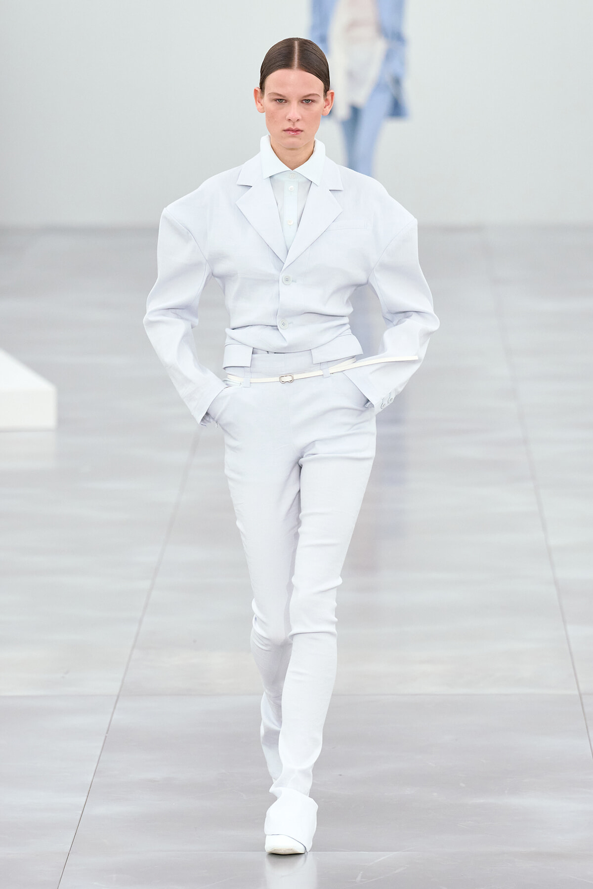 Model on the runway wearing a pale blue tailored suit with a belt, hands in pockets, neutral expression.