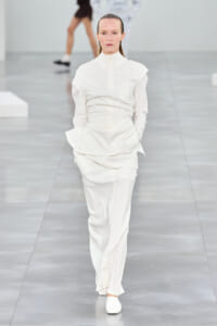 Female model walking a runway in an all-white, layered button-down outfit with matching pants and white shoes.