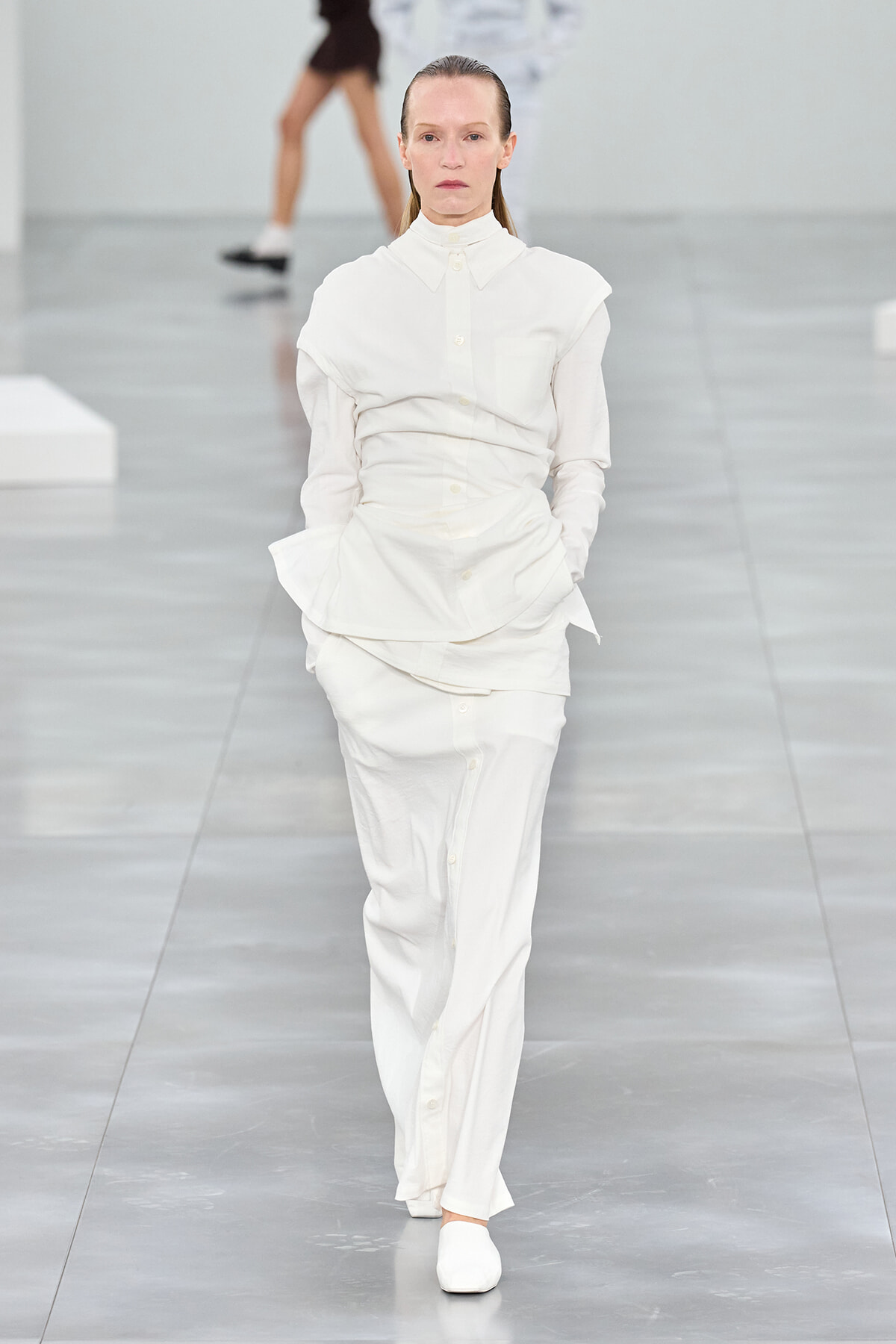 Female model walking a runway in an all-white, layered button-down outfit with matching pants and white shoes.