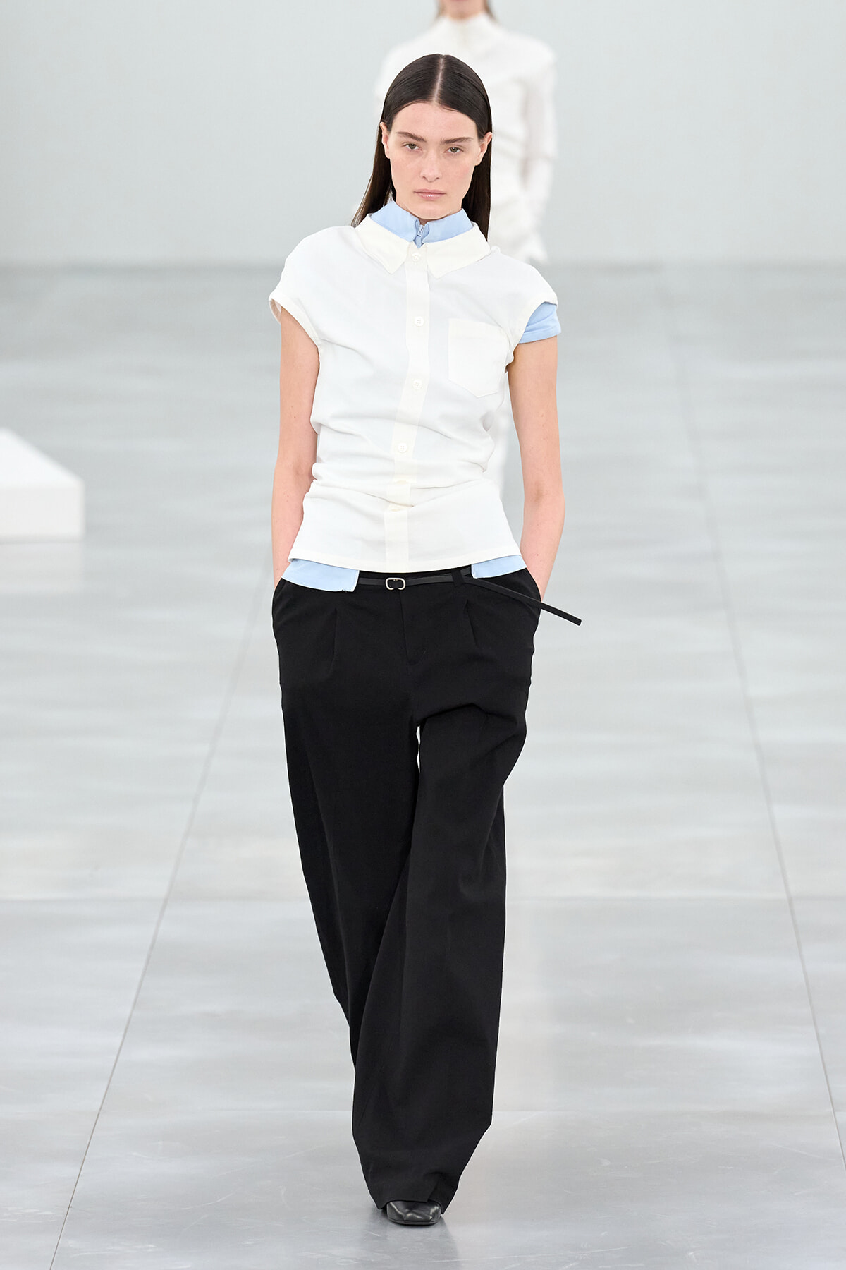 Model walks a runway in a white short-sleeve button-down shirt over a light blue layer, paired with black wide-leg trousers and a slim belt.