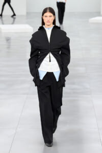 Model walks a minimalist runway in a black, oversized zip-front jacket over a white shirt with light blue accents and wide black pants.