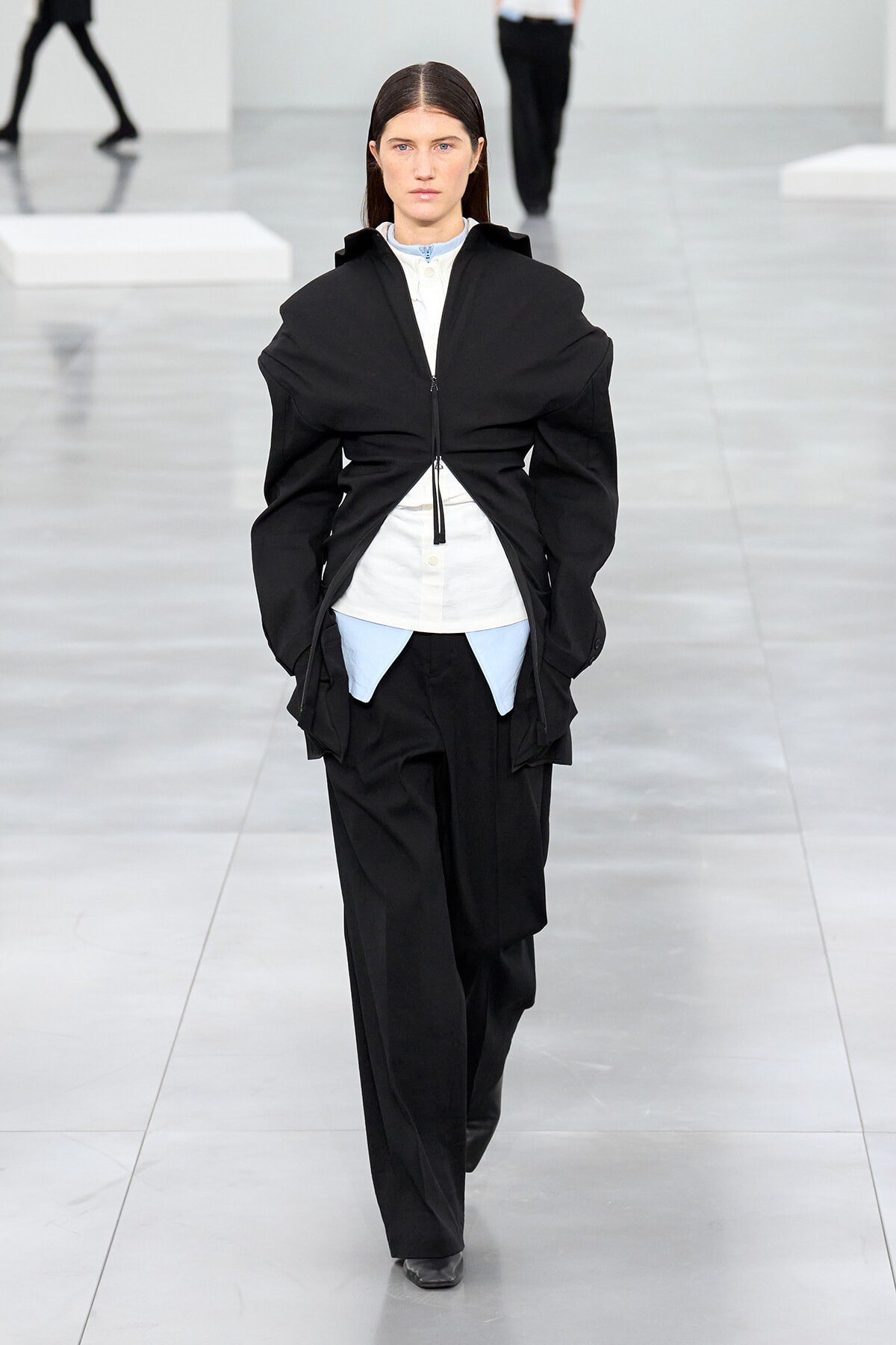 Model walks a minimalist runway in a black, oversized zip-front jacket over a white shirt with light blue accents and wide black pants.