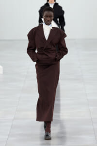 Runway model walking in a dark brown double-breasted suit with a white high-collar shirt and black shoes.