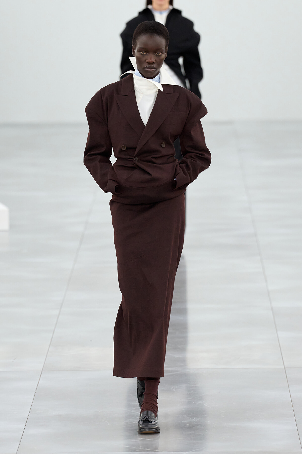Runway model walking in a dark brown double-breasted suit with a white high-collar shirt and black shoes.