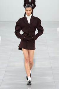 Model walks the runway wearing an oversized dark brown blazer with matching miniskirt, white shirt, and black loafers with white socks, hands in pockets.
