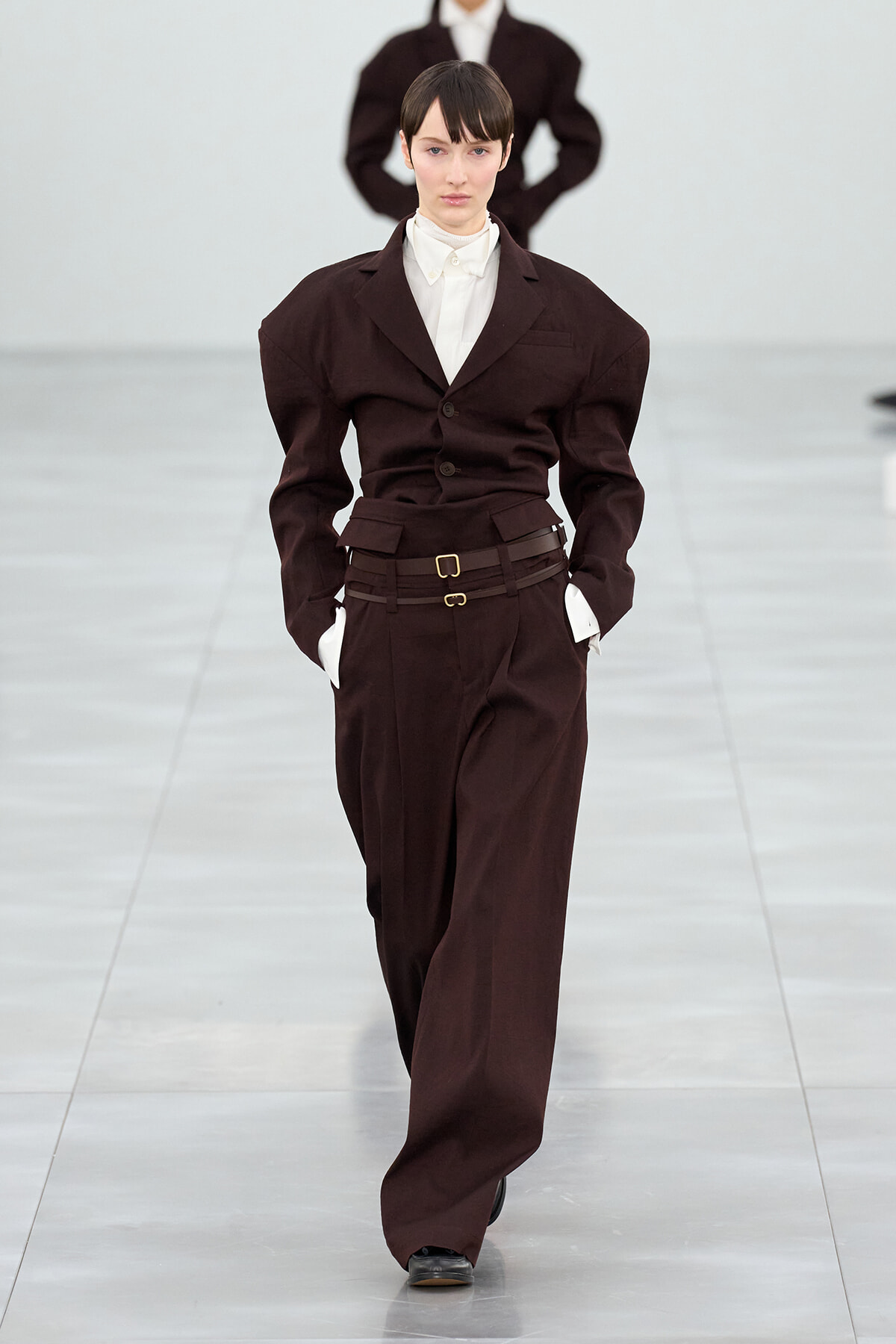 Model on a runway wearing a burgundy suit with a white shirt, hands in pockets, mid-stride.