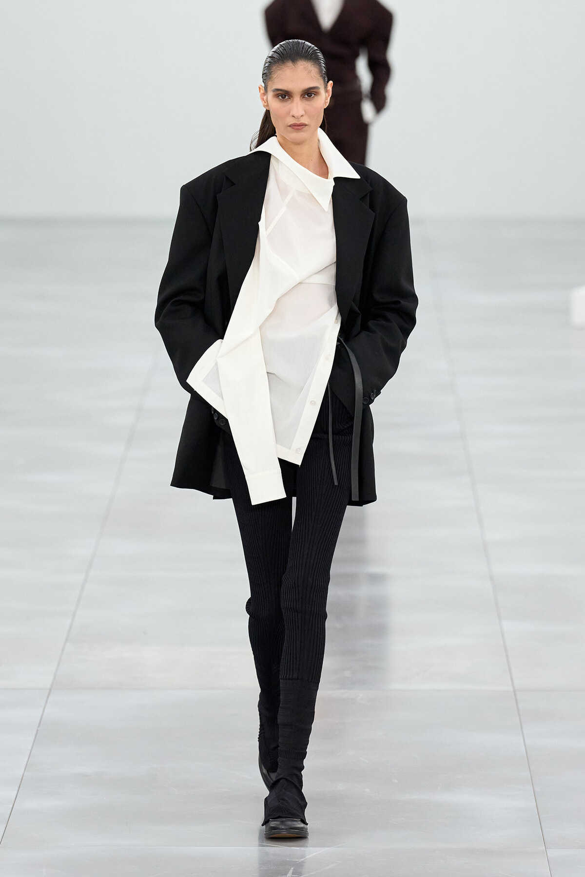 Model walks a minimalist runway in an oversized black blazer over a white layered shirt and black ribbed pants with black ankle boots.