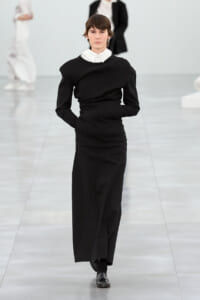 Model walking a minimalist black long dress with a white collared shirt visible at the neck on a fashion runway