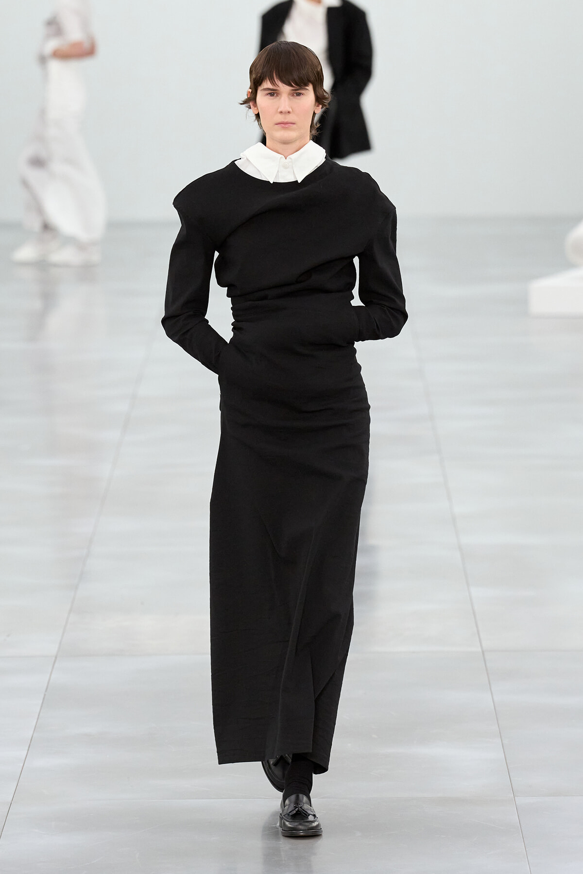 Model walking a minimalist black long dress with a white collared shirt visible at the neck on a fashion runway