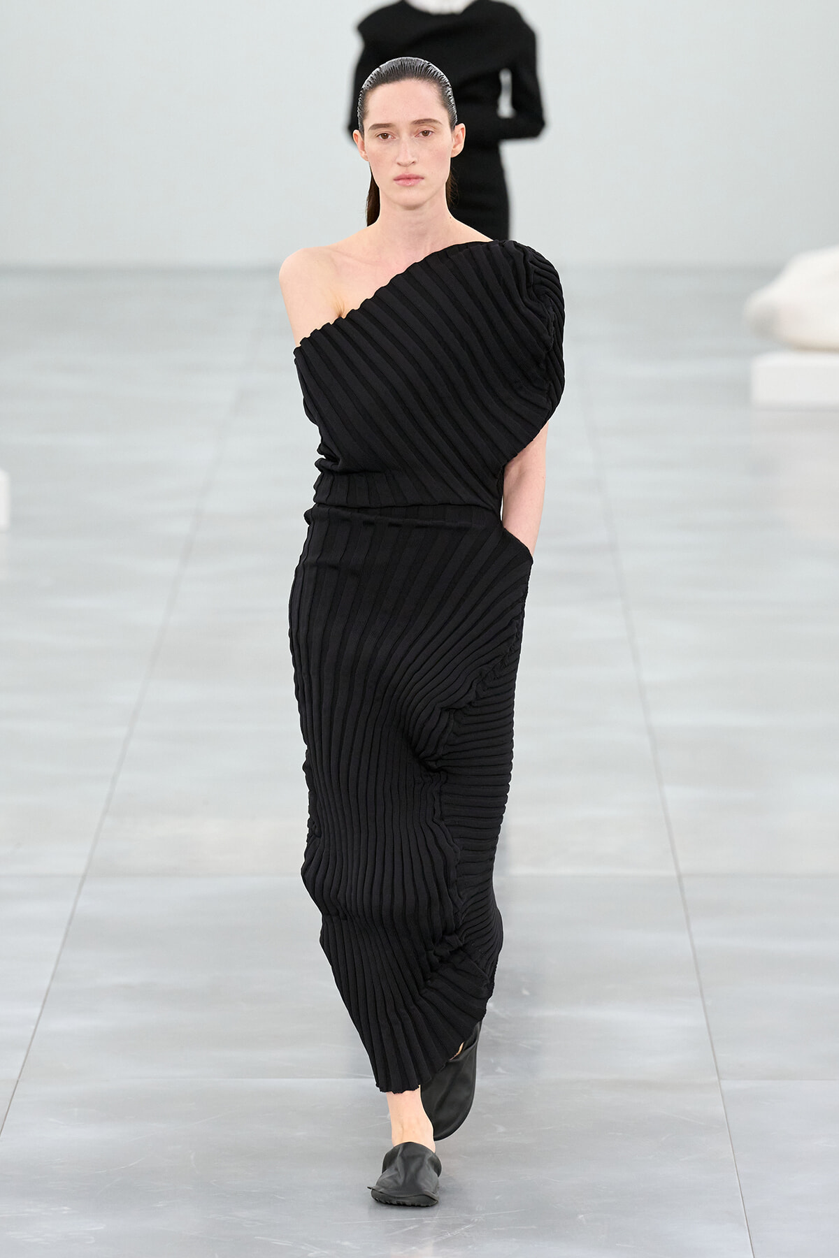 Model walking a runway in a full-length black pleated one-shoulder dress with diagonal pleats and black flats.