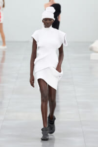 Female model walks the runway in a white ribbed knit dress with sculpted shoulders and matching turban, paired with gray ankle boots.