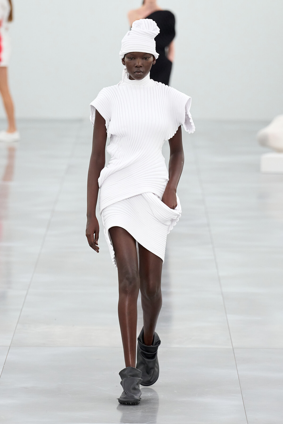 Female model walks the runway in a white ribbed knit dress with sculpted shoulders and matching turban, paired with gray ankle boots.