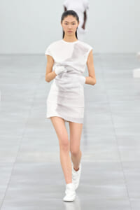 Asian model on a runway wearing a white textured mini dress and white boots, mid-stride.
