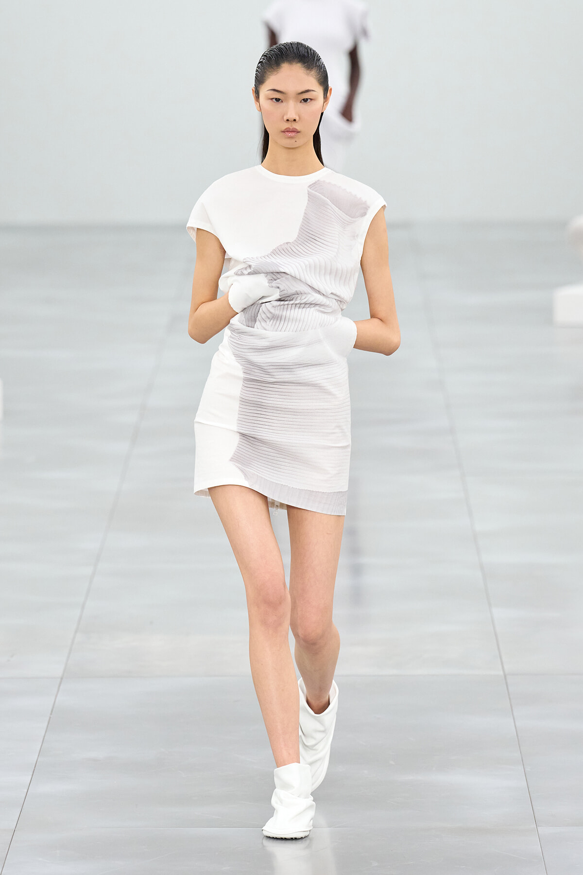Asian model on a runway wearing a white textured mini dress and white boots, mid-stride.