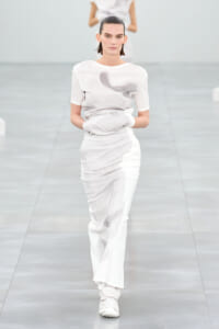 Female model walks a white-toned knit outfit on a minimalist runway, hands tucked in.