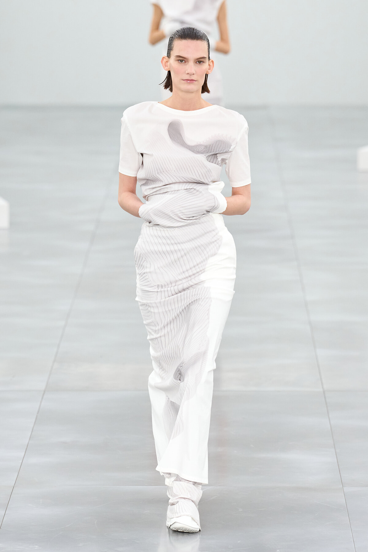 Female model walks a white-toned knit outfit on a minimalist runway, hands tucked in.