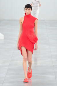 Model walks the runway in a bright red, ribbed, sleeveless high-neck dress with a sculpted drape at the hip and matching red slip-on shoes on a pale runway.