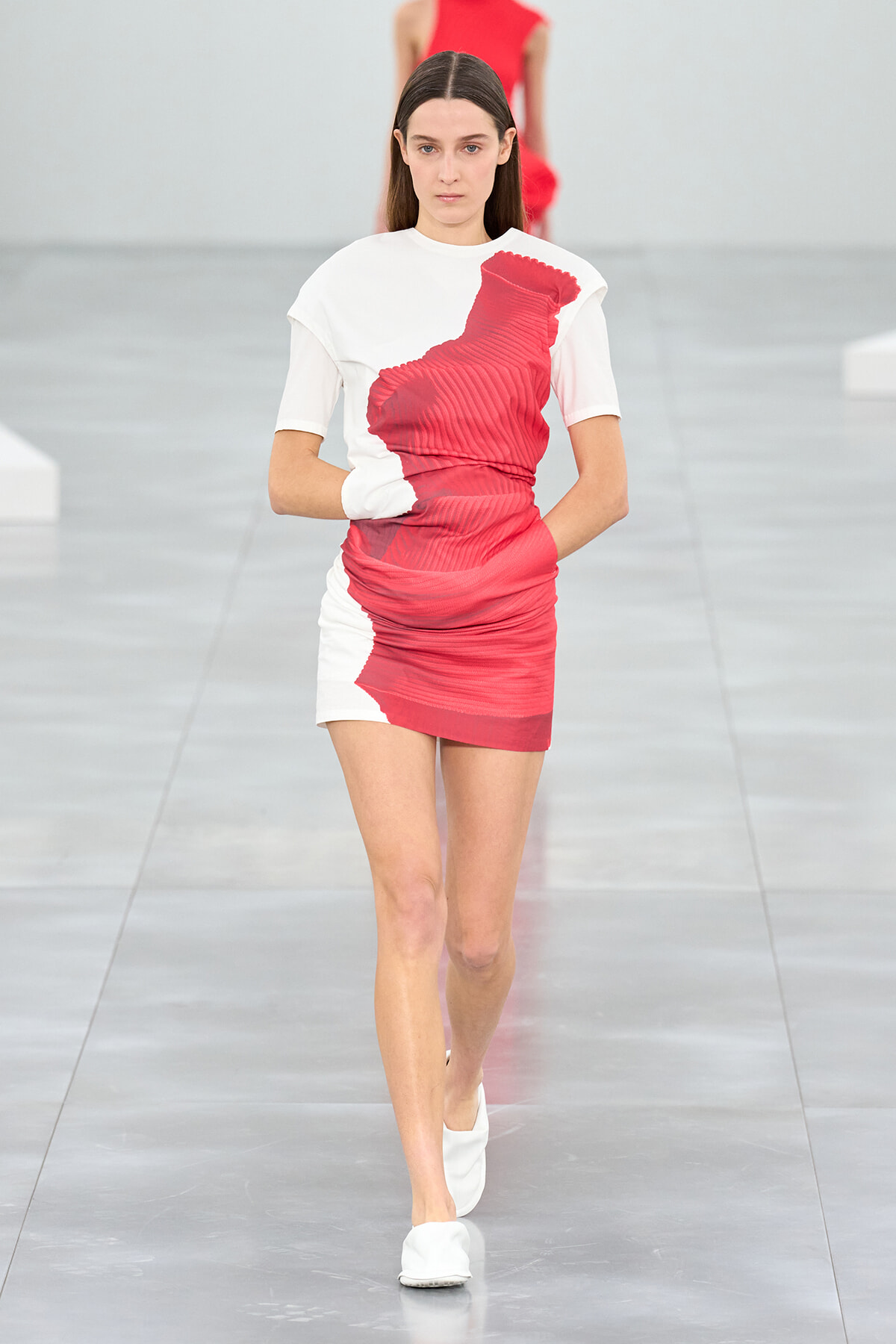 Model walking the runway in a white mini dress with a bold red abstract print, white slip-on shoes.
