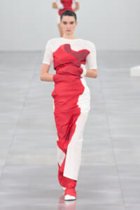 Model walking a white-and-red color-block dress with pleated red panels on a minimalist runway.