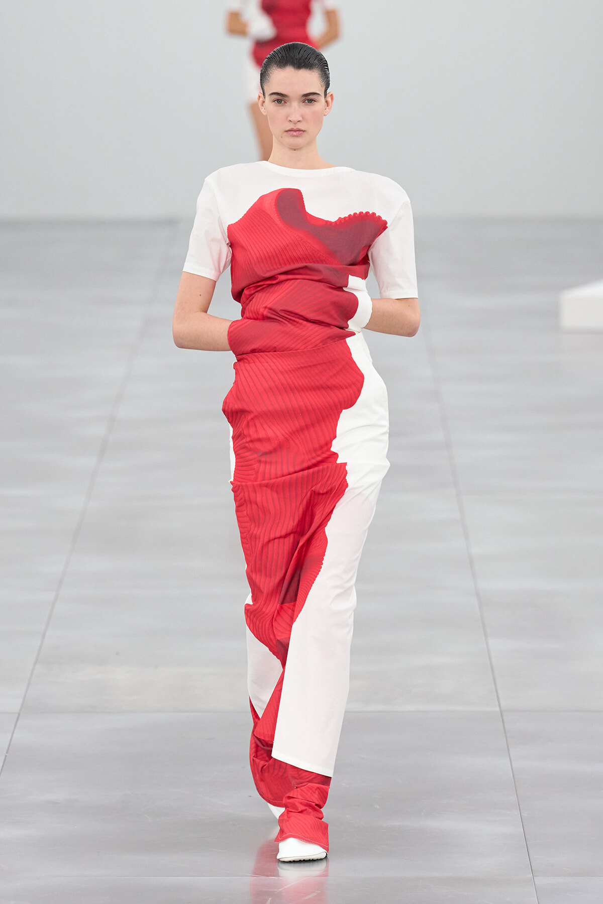Model walking a white-and-red color-block dress with pleated red panels on a minimalist runway.