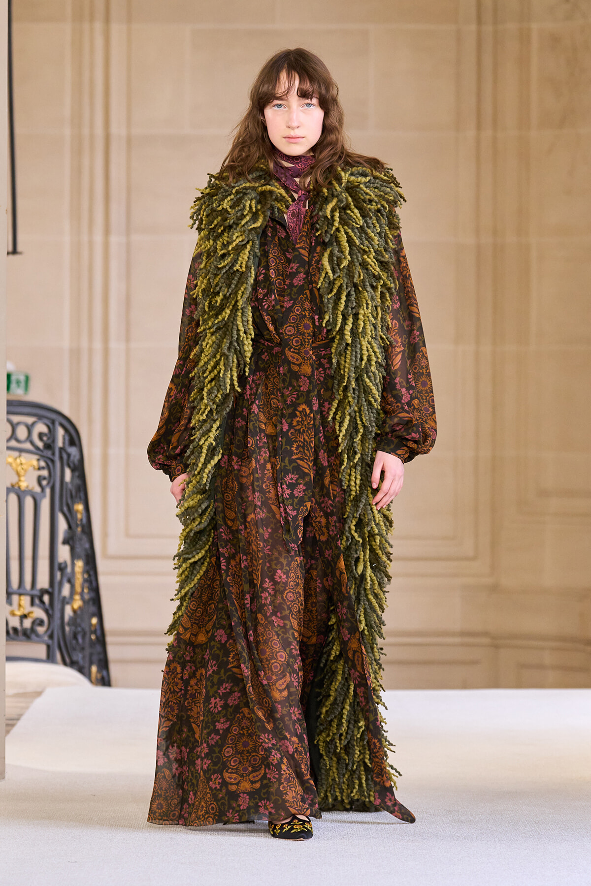 Model walks the runway in a long brown paisley dress with a large green shaggy scarf.