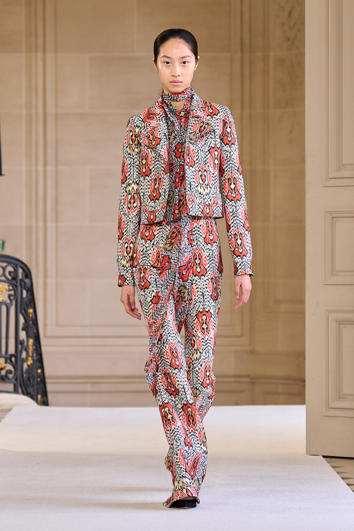 Runway model in a red, black, and white patterned suit with a matching scarf on a catwalk.