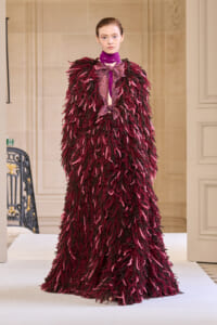 Model on a runway wearing a voluminous burgundy shaggy coat with pink accents and a purple neck scarf, haute couture look for a fashion show.