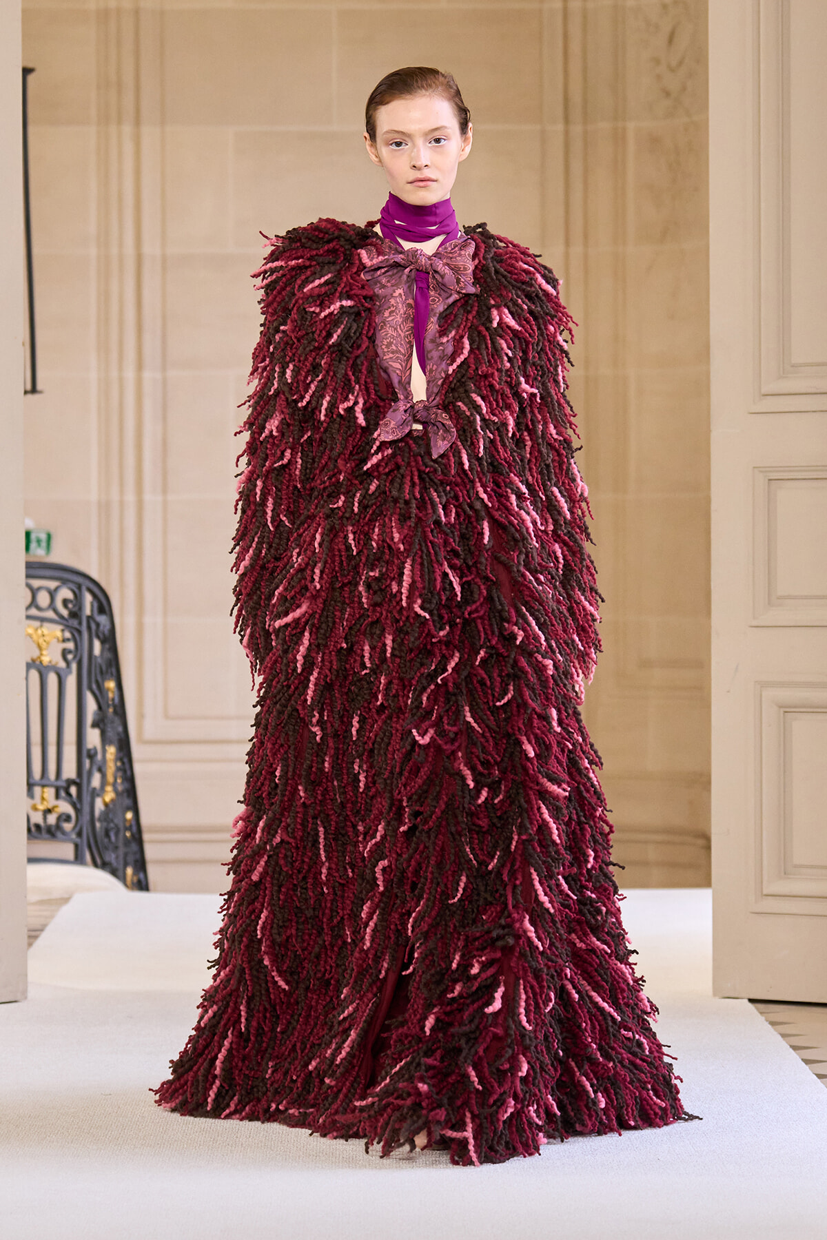 Model on a runway wearing a voluminous burgundy shaggy coat with pink accents and a purple neck scarf, haute couture look for a fashion show.