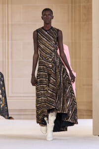 Model walking a runway in a sleeveless, gold-and-black sequined striped gown with an asymmetrical hem and white knee-high boots.