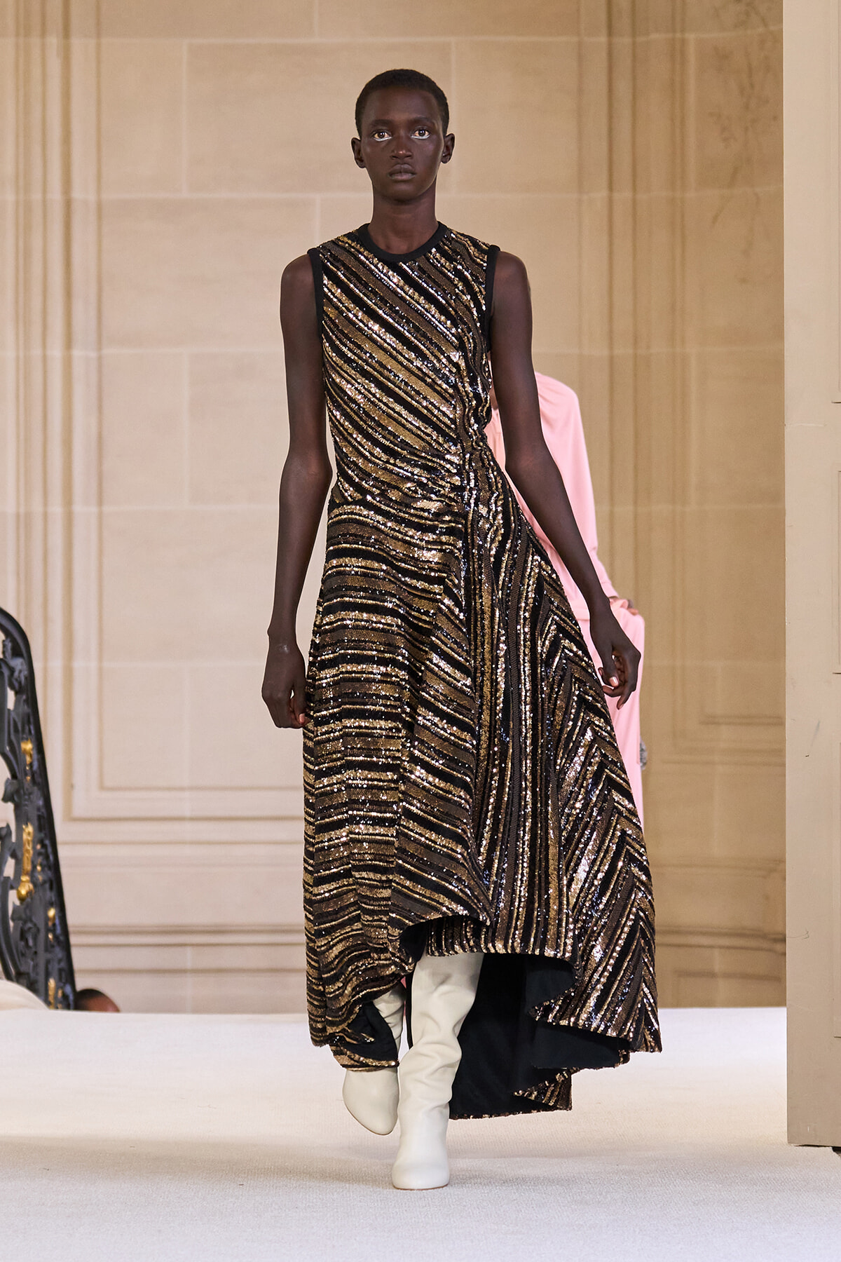 Model walking a runway in a sleeveless, gold-and-black sequined striped gown with an asymmetrical hem and white knee-high boots.