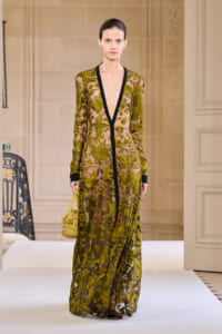 Model walking a runway in a sheer olive-green floral lace gown with black trim and deep neckline, carrying a woven yellow bag.