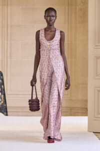 Model on a runway wearing a pink textured sleeveless jumpsuit with a belt and gold buttons, carrying a woven purple handbag.