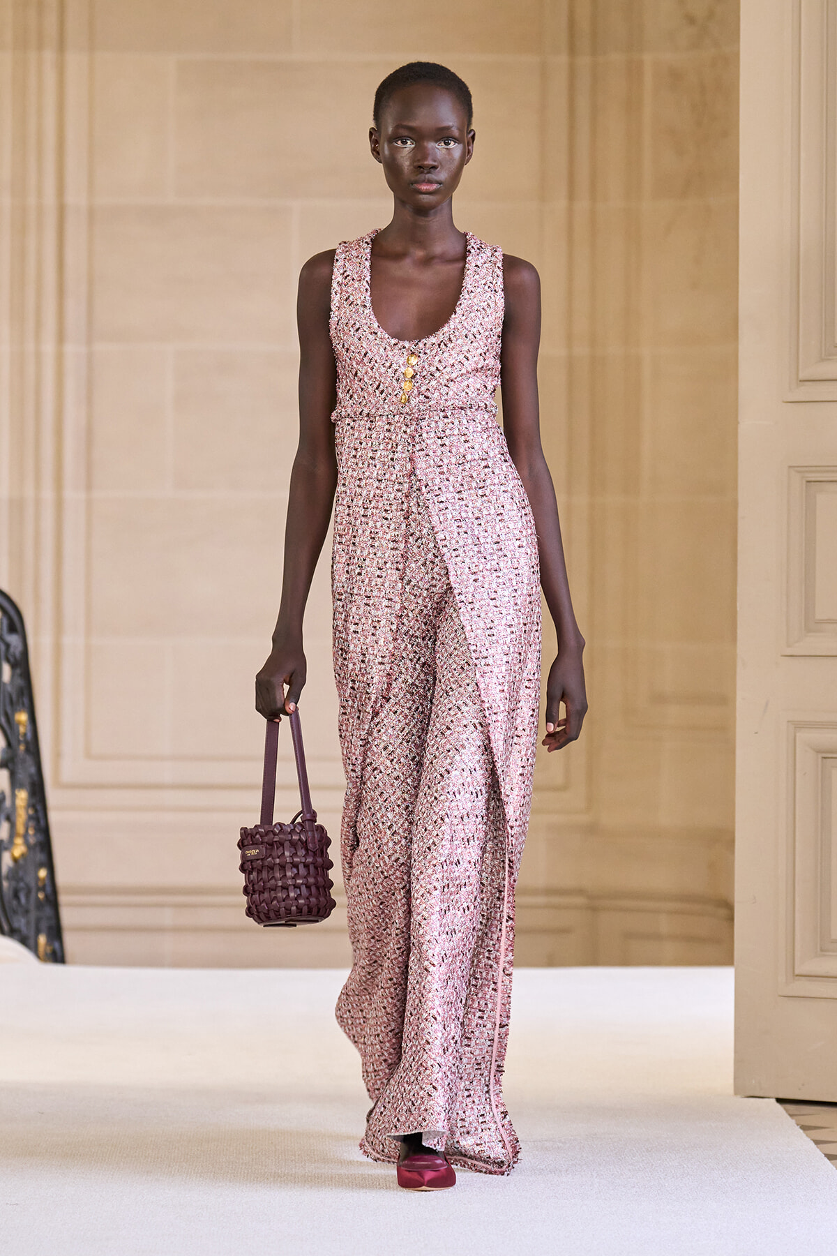 Model on a runway wearing a pink textured sleeveless jumpsuit with a belt and gold buttons, carrying a woven purple handbag.