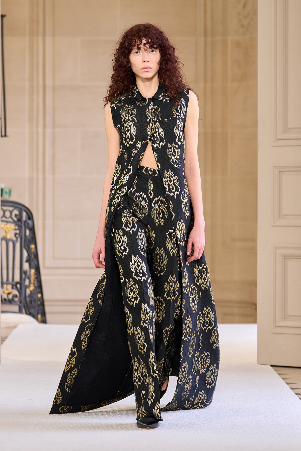 Female model walking a runway in a black and gold patterned sleeveless outfit with a draped cape.